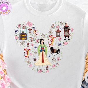 May include: White t-shirt featuring a colorful graphic of Mulan and other characters arranged in a Mickey Mouse head shape. The design includes pink flowers, a red dragon, and a black horse. The shirt has a crew neck and short sleeves.