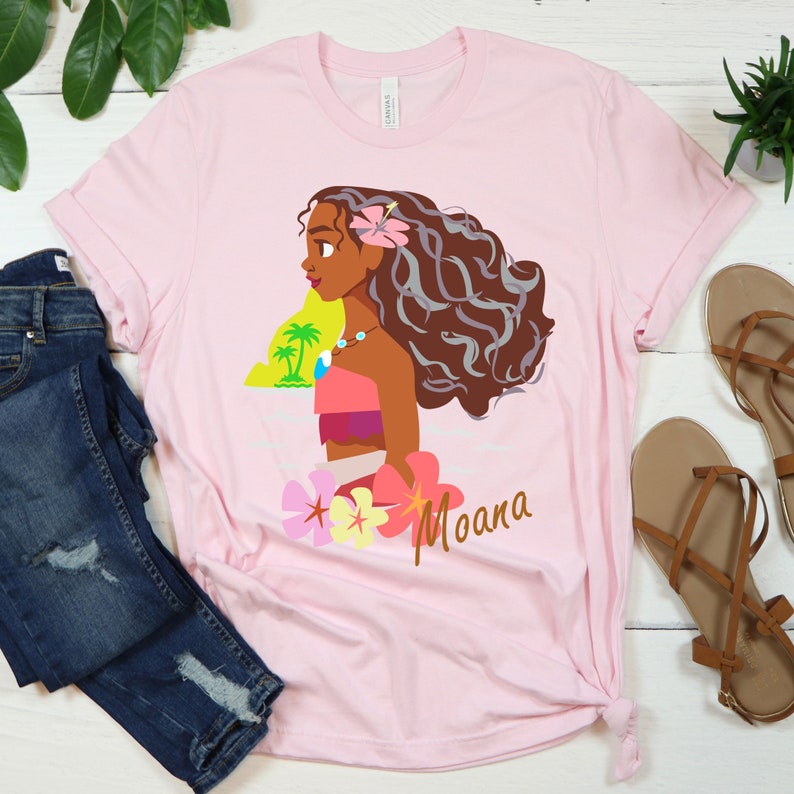 May include: Pink t-shirt with a cartoon image of Moana, a Polynesian princess from the Disney movie Moana. The image shows Moana with long brown hair, a flower in her hair, and a necklace. The text "Moana" is printed below the image.