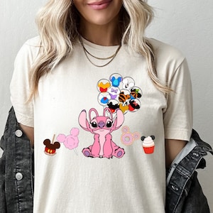 May include: A light beige t-shirt with a pink Stitch character holding a bunch of colorful balloons with Disney characters. The balloons are Mickey Mouse, Minnie Mouse, Donald Duck, Goofy, Pluto, and others. There are also Mickey Mouse themed treats on the bottom of the shirt, including a Mickey Mouse head pretzel, cotton candy, a donut, and a cupcake.