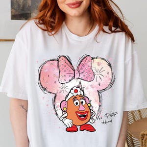 May include: White t-shirt with a pink and white watercolor design of a Mickey Mouse head with a pink bow. The design also includes a cartoon image of Mrs. Potato Head with the text "Mrs. Potato Head" below.