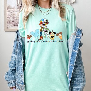 May include: A light green t-shirt with a graphic of Woody and Buzz Lightyear from Toy Story, Mickey Mouse ears, and other Disney characters. The text "BEST DAY EVER" is printed below the graphic.