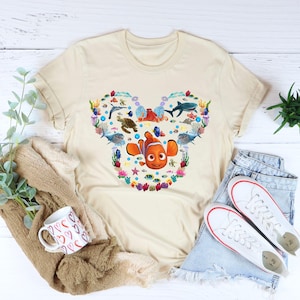 May include: Beige t-shirt featuring a heart-shaped design with cartoon sea creatures, including Nemo. The design includes fish, sharks, and coral. The shirt is paired with denim shorts and white sneakers.