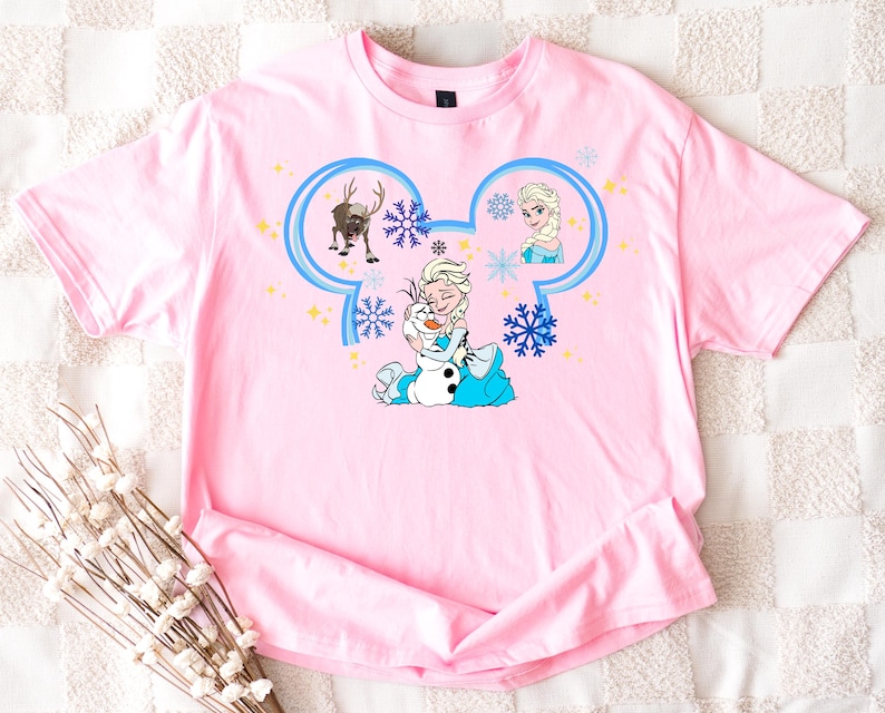 A pink t-shirt with a cartoon design featuring characters from the Disney movie Frozen, including Elsa, Olaf, and other beloved characters.
