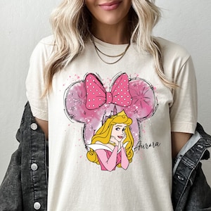 May include: A white t-shirt with a pink and white cartoon image of Sleeping Beauty with a pink bow in the shape of Mickey Mouse ears. The text "Aurora" is written in pink below the image.