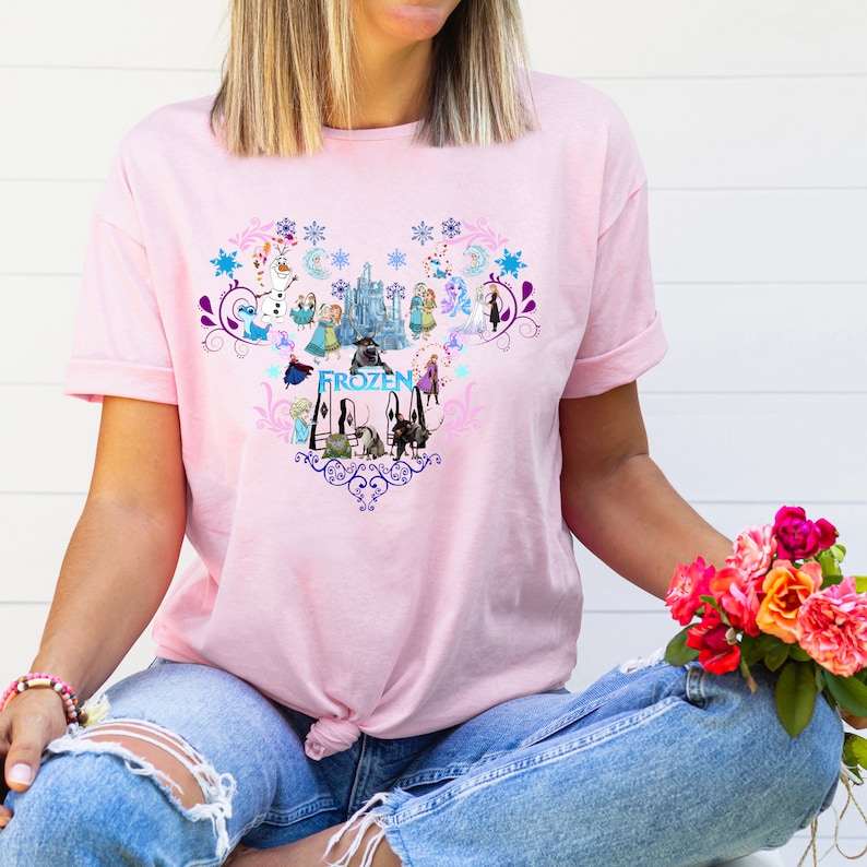 Puede incluir: Camiseta rosa con un dise&ntilde;o en forma de coraz&oacute;n con personajes de la pel&iacute;cula Frozen. El dise&ntilde;o incluye a Elsa, Anna, Olaf, Kristoff, Sven y el castillo. La palabra "Frozen" est&aacute; escrita en letras azules.