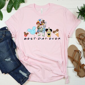 Puede incluir: Camiseta rosa con un gr&aacute;fico de personajes de Disney, incluyendo Mickey Mouse, Minnie Mouse, Woody, Buzz Lightyear y otros. El texto "BEST DAY EVER" est&aacute; impreso debajo del gr&aacute;fico.