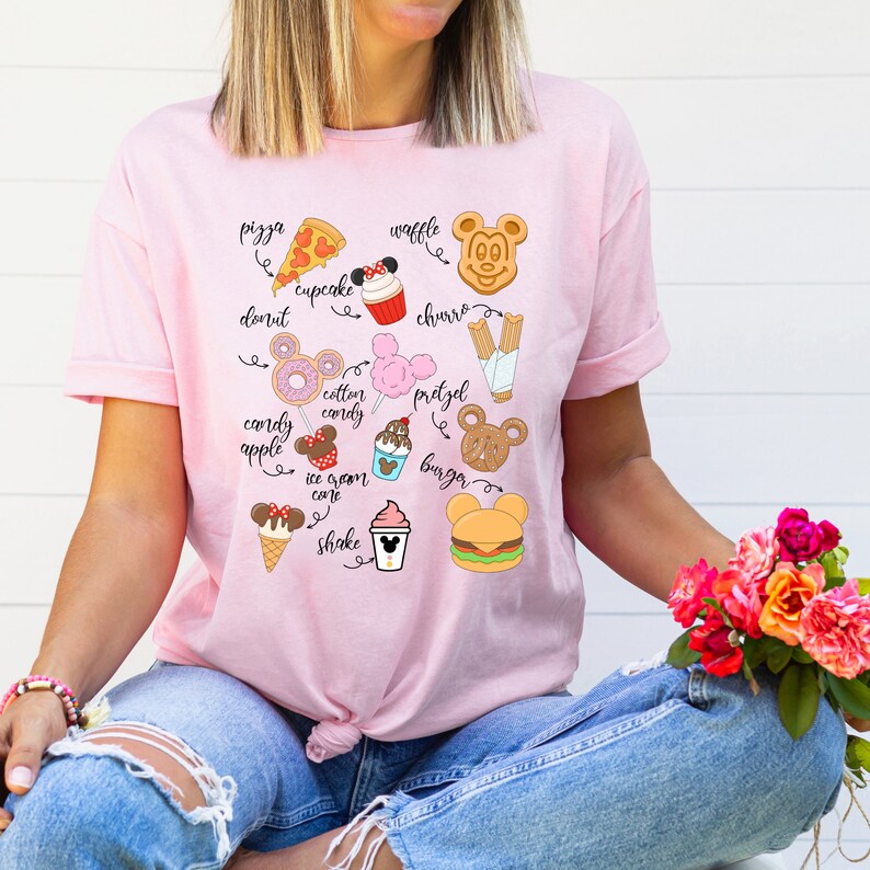 May include: Pink t-shirt with a graphic of various Disney park food items, including a Mickey Mouse waffle, a pretzel, a churro, a cupcake, a pizza slice, a donut, a cotton candy, a candy apple, an ice cream cone, a shake, and a burger. The text on the shirt reads "pizza", "waffle", "churro", "donut", "cotton candy", "pretzel", "candy apple", "ice cream cone", "shake", and "burger".