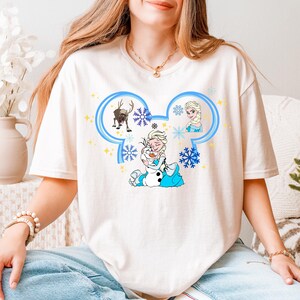 A woman wearing a white t-shirt with a Disney character design, including Elsa, Olaf, and a snowman, sitting on a couch.