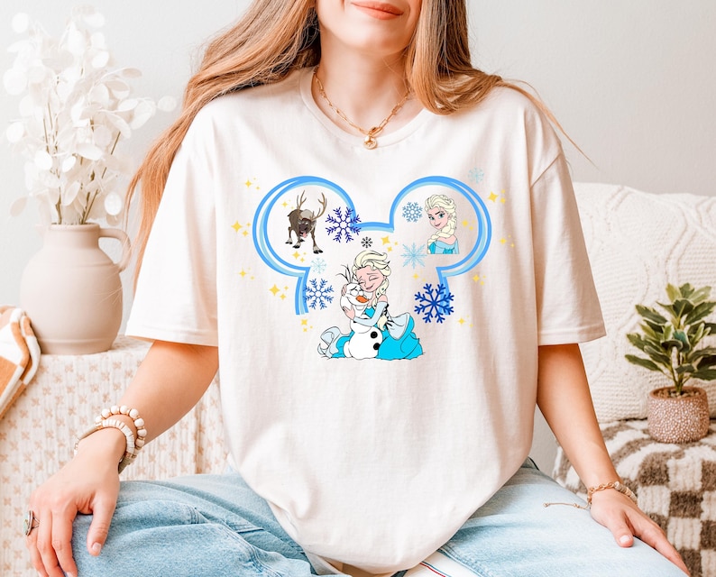 A woman wearing a white t-shirt with a Disney character design, including Elsa, Olaf, and a snowman, sitting on a couch.