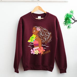 May include: A maroon sweatshirt with a graphic print of Moana, a Disney princess, standing in the ocean with a flower in her hair. The text "Moana" is printed below the image.