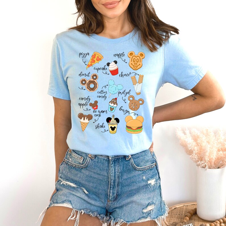 May include: Light blue t-shirt with a graphic of various Disney park food items, including Mickey Mouse ears, a pretzel, a cupcake, a waffle, a churro, a cotton candy, a burger, a corn dog, an ice cream cone, a shake, a pizza, a donut, and an apple. The text on the shirt reads "pizza", "cupcake", "donut", "cotton candy", "pretzel", "burger", "ice cream cone", "shake", "waffle", "churro", "apple", "cordy", "cordy".