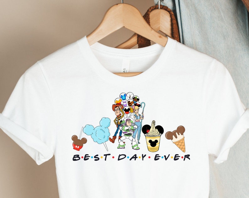 Puede incluir: Camiseta blanca con un gr&aacute;fico de personajes de Toy Story, incluyendo Woody, Buzz Lightyear y Jessie, sosteniendo globos y orejas de Mickey Mouse. El texto "BEST. DAY. EVER." est&aacute; impreso debajo del gr&aacute;fico.