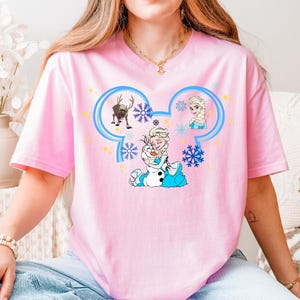 A woman wearing a pink t-shirt with a Disney character design, including Elsa, Olaf, and other characters from the Disney movie Frozen.