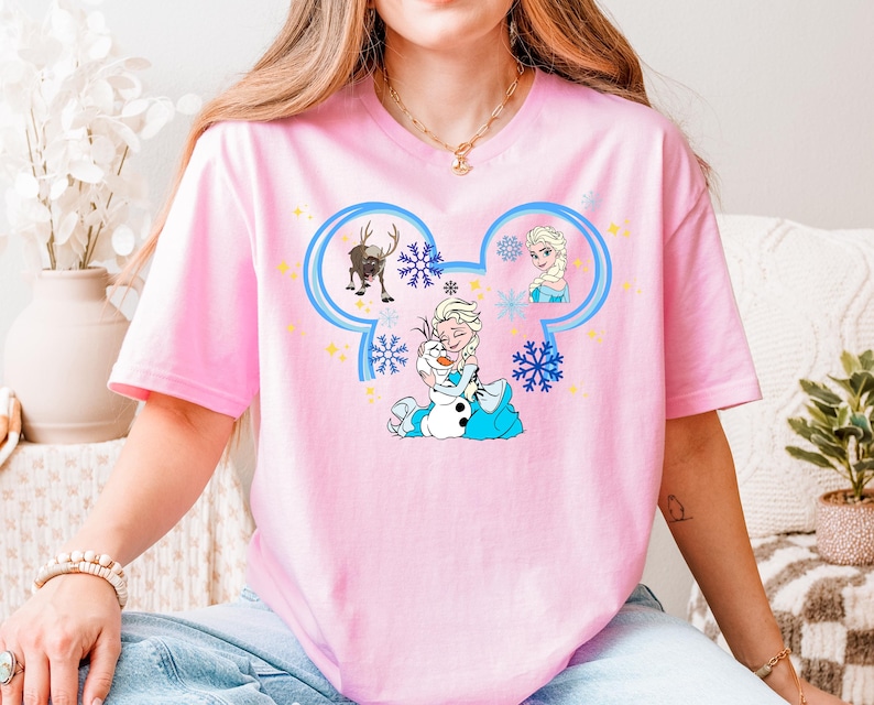 A woman wearing a pink t-shirt with a Disney character design, including Elsa, Olaf, and other characters from the Disney movie Frozen.
