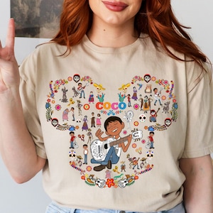 May include: A beige t-shirt with a colorful graphic of characters from the Disney Pixar movie "Coco". The graphic features Miguel playing a guitar and surrounded by other characters from the film. The word "Coco" is written in a colorful font.