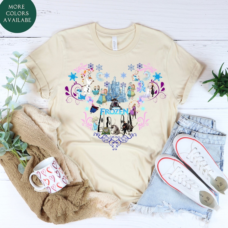 Puede incluir: Una camiseta beige con un dise&ntilde;o en forma de coraz&oacute;n que presenta personajes de la pel&iacute;cula Frozen. El dise&ntilde;o incluye a Elsa, Anna, Olaf, Kristoff, Sven y el castillo. El texto "FROZEN" est&aacute; escrito en letras azules.