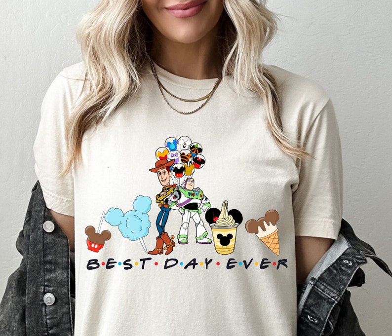 May include: A beige t-shirt with a graphic of Woody and Buzz Lightyear from Toy Story holding balloons. The graphic also includes Mickey Mouse ears, a Mickey Mouse ice cream cone, and a Mickey Mouse pretzel. The text "BEST DAY EVER" is printed below the graphic.