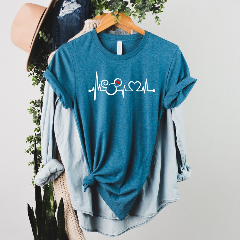 May include: A teal blue t-shirt with a white Mickey Mouse head and a red heart inside the ear, with a white heartbeat line on either side of the Mickey Mouse head.