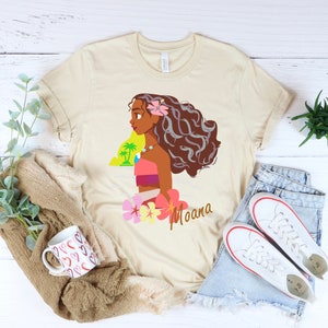 May include: A beige t-shirt with a cartoon image of Moana from the Disney movie Moana. The image shows Moana with her long brown hair, wearing a pink and orange outfit, and holding a flower. The text "Moana" is printed below the image.
