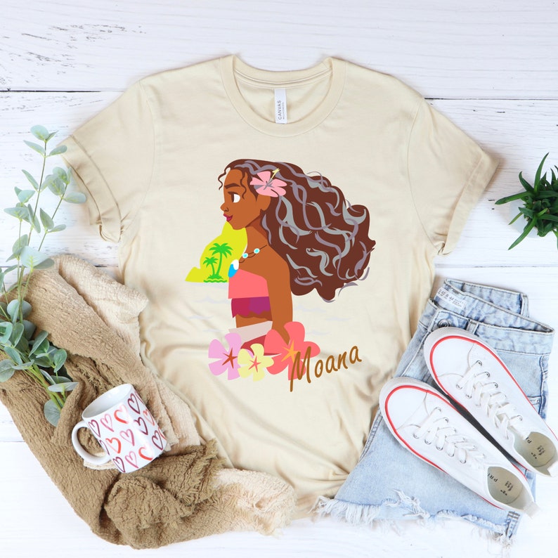 May include: A beige t-shirt with a cartoon image of Moana from the Disney movie Moana. The image shows Moana with her long brown hair, wearing a pink and orange outfit, and holding a flower. The text "Moana" is printed below the image.