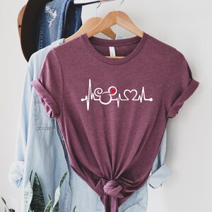 May include: A maroon t-shirt with a white graphic of a heartbeat with a Mickey Mouse head and a heart in the middle.