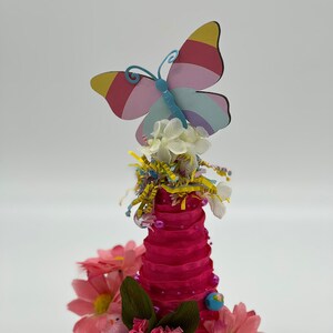 Op de afbeelding: Een grillig decoratief stuk met een roze kegelvormige structuur versierd met roze bloemen, groene bladeren en een kleurrijke vlinder. De basis is een witte theekop, wat een charmante, artistieke weergave creëert.