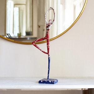 May include: A decorative metal sculpture of a ballerina in a graceful pose. The sculpture features red, white, and blue accents, standing on a blue base. A gold-framed round mirror hangs on the wall behind the sculpture.