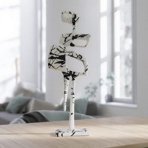 May include: Abstract sculpture with a white base and a vertical structure. The sculpture features black and white patterned shapes, resembling a stylized figure. It stands on a light wood table in a bright, modern interior setting.