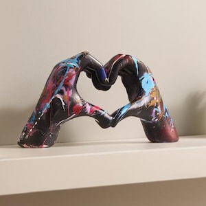May include: A decorative sculpture of two hands forming a heart shape. The sculpture is painted with a colorful graffiti-style design. It sits on a white shelf with books and a small potted plant.