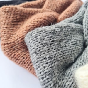 May include: Close-up of several knitted headbands in various colours. The headbands are made of a thick, textured yarn. Colours include shades of brown, grey, and off-white. The knit pattern is visible, creating a cosy and warm appearance.