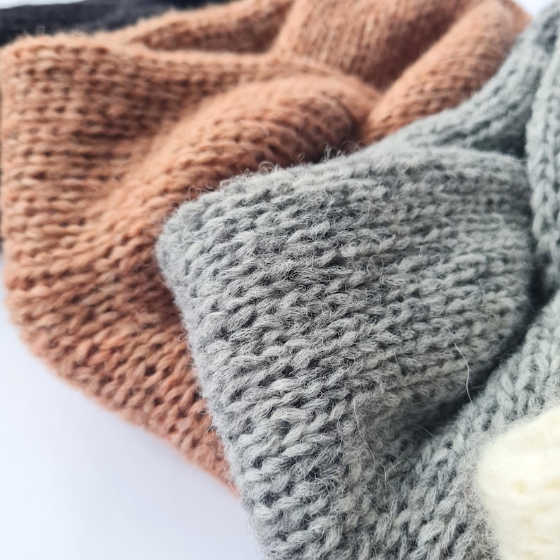 May include: Close-up of several knitted headbands in various colours. The headbands are made of a thick, textured yarn. Colours include shades of brown, grey, and off-white. The knit pattern is visible, creating a cosy and warm appearance.