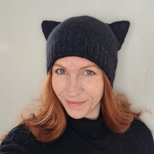 Peut inclure: Un bonnet en tricot gris foncé avec des oreilles de chat. Le chapeau est fait d'une matière douce et texturée. Le chapeau est porté par une personne aux cheveux roux et un col roulé noir.