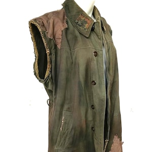 Puede incluir: Chaqueta sin mangas verde oliva con parches marrones en los hombros y las mangas. La chaqueta tiene una botonadura frontal, un cuello y un bolsillo. La tela parece desgastada con costuras y desgaste visibles.