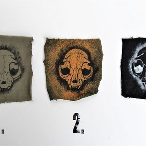 May include: Three fabric squares with a black and white cat skull design printed on them. The squares are numbered 1, 2, and 3. The fabric on square 1 is a dark green color, the fabric on square 2 is a light orange color, and the fabric on square 3 is a black color.