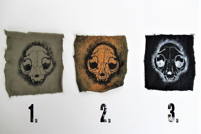 May include: Three fabric squares with a black and white cat skull design printed on them. The squares are numbered 1, 2, and 3. The fabric on square 1 is a dark green color, the fabric on square 2 is a light orange color, and the fabric on square 3 is a black color.