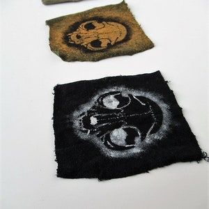 May include: Three fabric patches with a skull design. The first patch is green with a black skull, the second is brown with a black skull, and the third is black with a white skull.