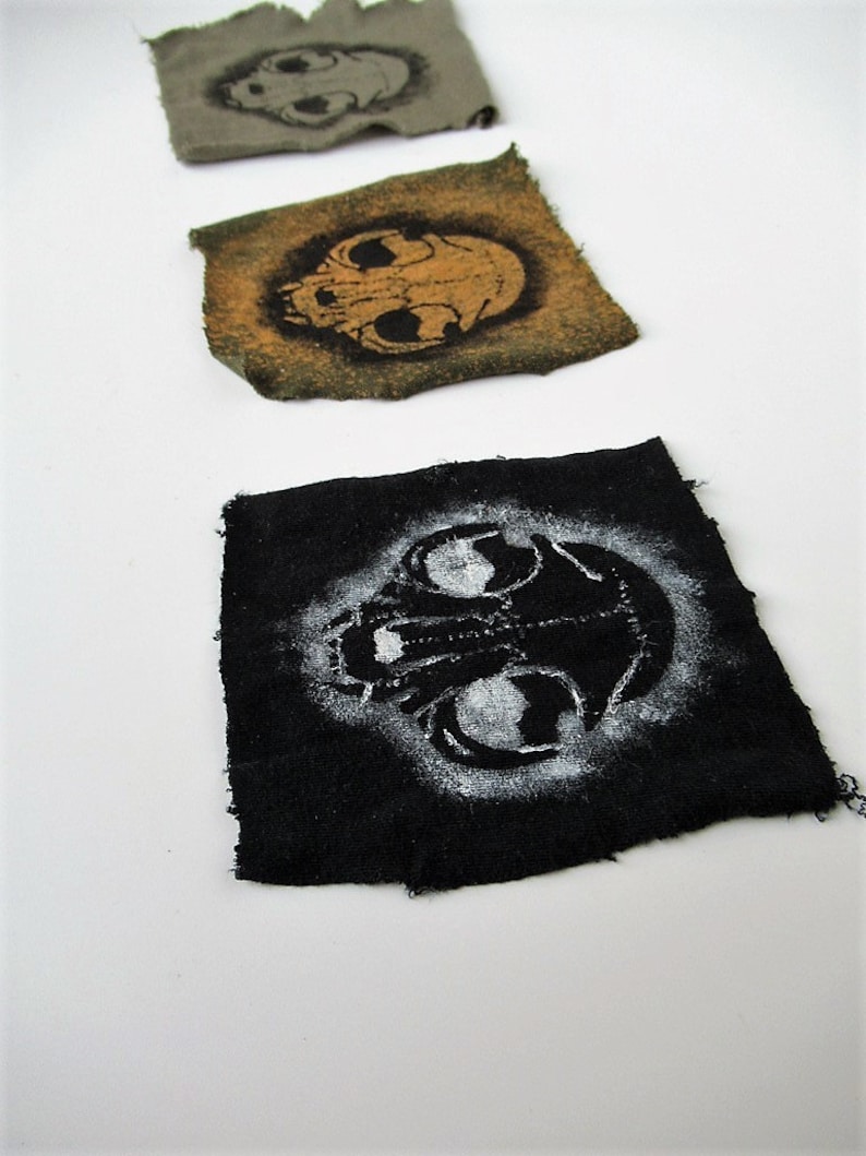 May include: Three fabric patches with a skull design. The first patch is green with a black skull, the second is brown with a black skull, and the third is black with a white skull.