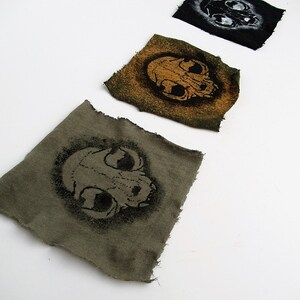 May include: Three fabric patches with a skull design. The patches are in different colors: black, green, and brown. The skull on the black patch is white, the skull on the green patch is orange, and the skull on the brown patch is black.
