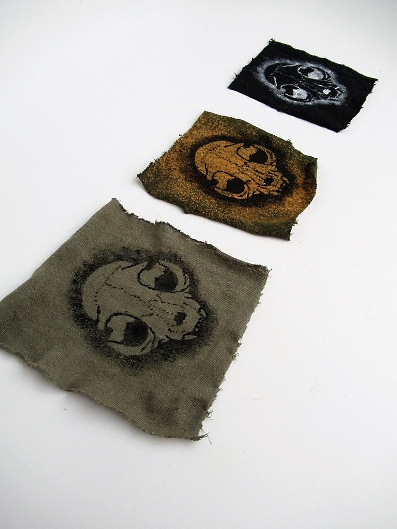 May include: Three fabric patches with a skull design. The patches are in different colors: black, green, and brown. The skull on the black patch is white, the skull on the green patch is orange, and the skull on the brown patch is black.