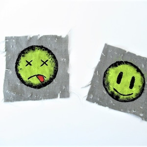 Op de afbeelding: Twee vierkante stukken stof met handgeschilderde smiley-gezichten. Eén gezicht is groen met zwarte ogen en een rode tong die uitsteekt. Het andere gezicht is groen met zwarte ogen en een zwarte glimlach.
