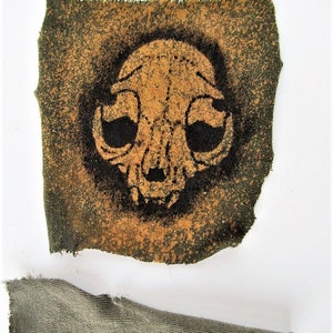 May include: A small, square patch of fabric with a black and orange skull design. The fabric is a dark green color with a rough texture.