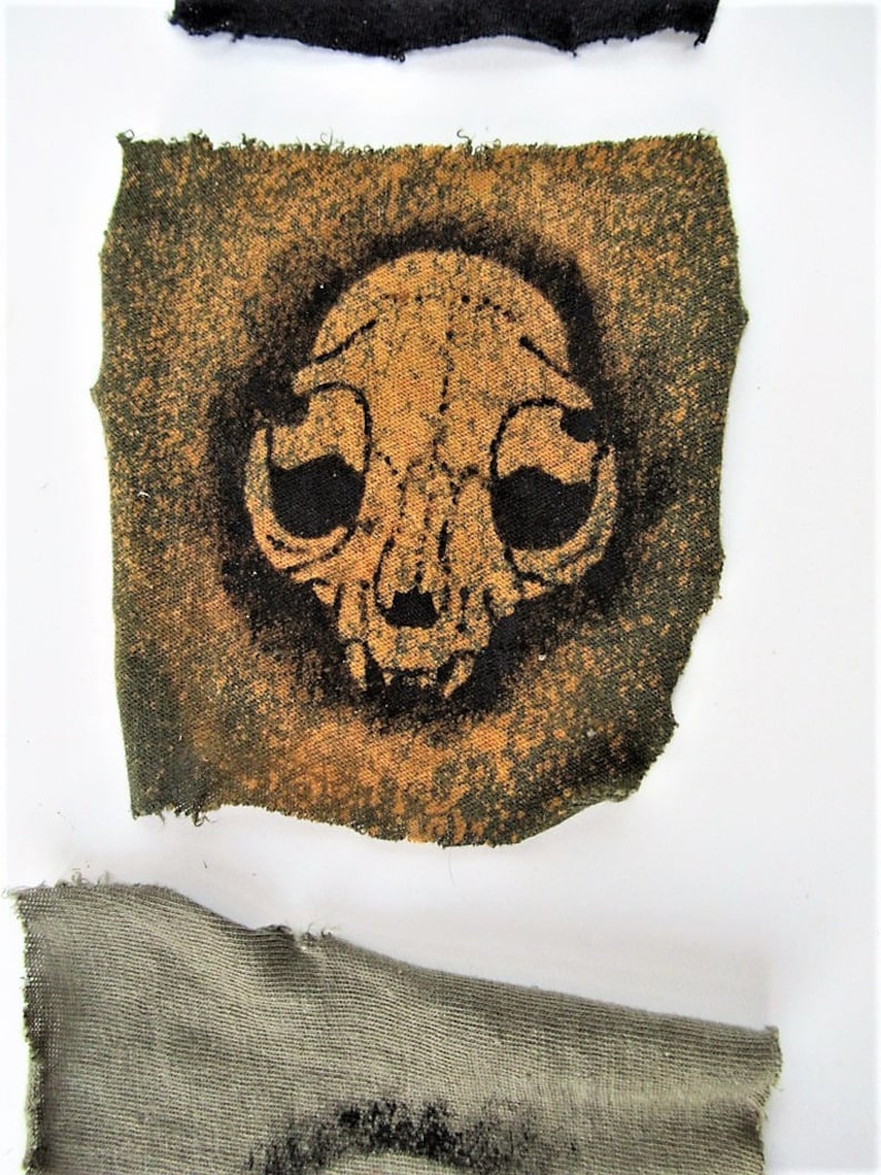 May include: A small, square patch of fabric with a black and orange skull design. The fabric is a dark green color with a rough texture.
