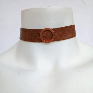 Puede incluir: Un collar gargantilla de cuero marrón con una hebilla circular. El collar se muestra en un maniquí blanco. El cuero parece tener textura y la hebilla es del mismo color que el collar.