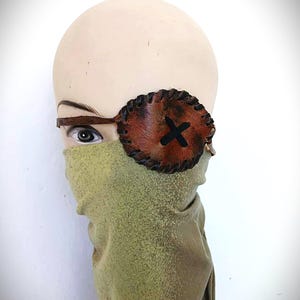 May include: A brown leather eye patch with a black cross design, worn on a mannequin head. The eye patch is stitched and has a dark brown leather strap. A green fabric covers the lower face.