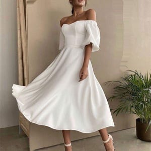 Puede incluir: Un vestido blanco con hombros descubiertos con un corpi&ntilde;o ajustado y una falda fluida. El vestido presenta mangas cortas y abullonadas y una cintura ce&ntilde;ida. La modelo lleva tacones blancos a juego. El vestido llega a la rodilla y parece estar hecho de una tela suave.