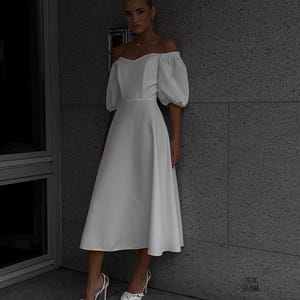 Puede incluir: Un vestido midi blanco con hombros descubiertos y mangas cortas abullonadas. El vestido tiene un corpi&ntilde;o ajustado y una falda acampanada. La modelo lleva tacones altos blancos. El fondo es una pared gris.