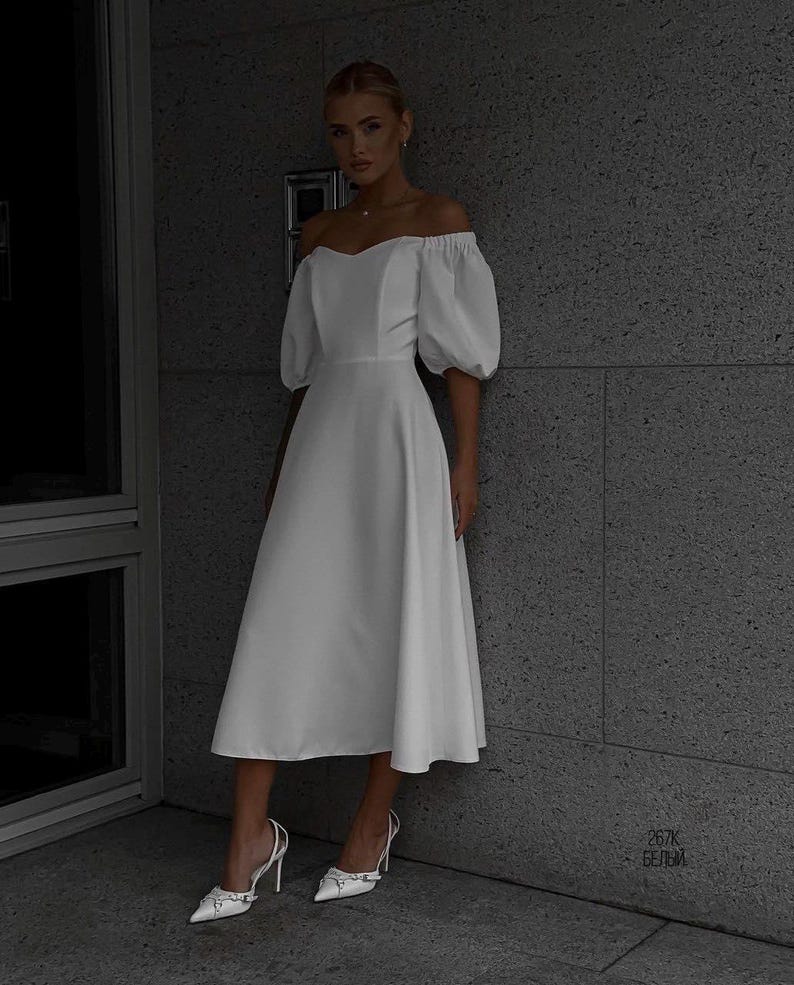 Puede incluir: Un vestido midi blanco con hombros descubiertos y mangas cortas abullonadas. El vestido tiene un corpi&ntilde;o ajustado y una falda acampanada. La modelo lleva tacones altos blancos. El fondo es una pared gris.