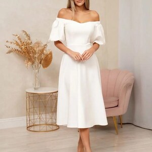 Puede incluir: Un vestido blanco con hombros descubiertos, mangas cortas abullonadas y cintura entallada. El vestido llega a la pantorrilla y se combina con tacones blancos de punta. El fondo incluye una mesa auxiliar dorada, un sill&oacute;n rosa y un jarr&oacute;n con flores secas.