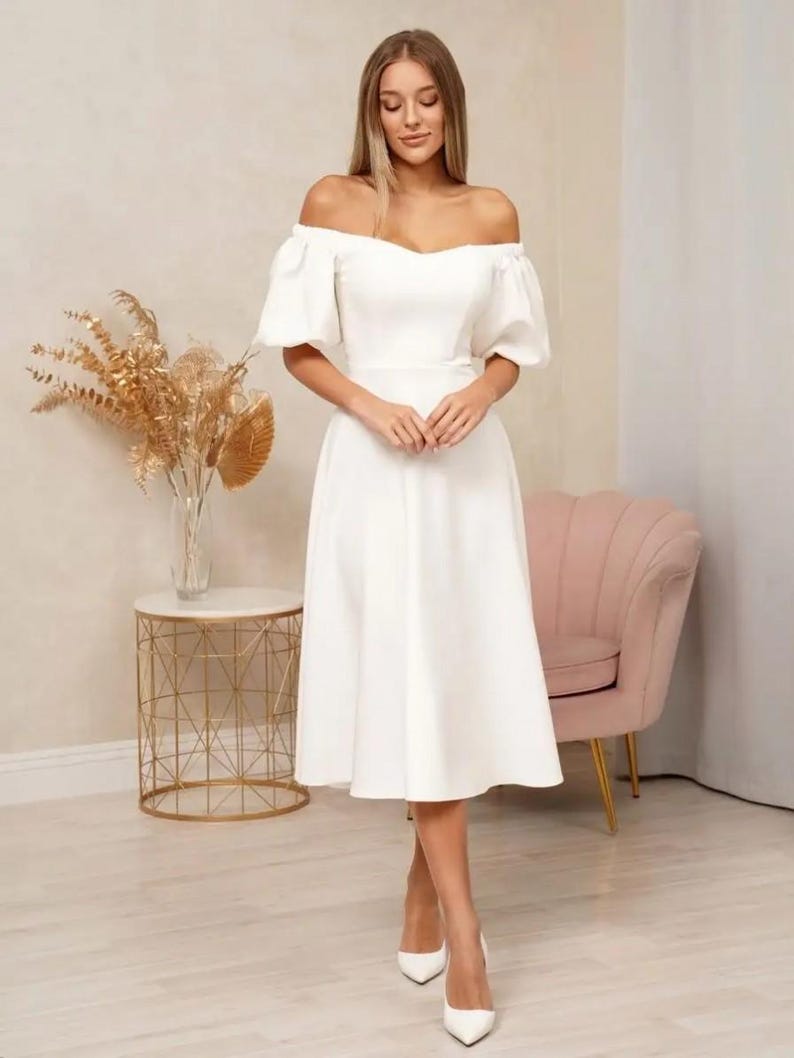 Puede incluir: Un vestido blanco con hombros descubiertos, mangas cortas abullonadas y cintura entallada. El vestido llega a la pantorrilla y se combina con tacones blancos de punta. El fondo incluye una mesa auxiliar dorada, un sill&oacute;n rosa y un jarr&oacute;n con flores secas.