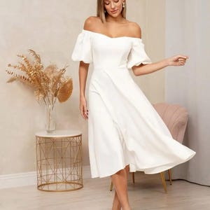Puede incluir: Un vestido blanco con hombros descubiertos y mangas cortas abullonadas. El vestido tiene un corpi&ntilde;o ajustado y una falda fluida que llega hasta la pantorrilla. La modelo lleva tacones blancos de punta. Una mesa auxiliar dorada y una silla rosa est&aacute;n en el fondo.